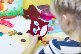 Tyttö ompelee kultalangalla huopakankaasta tekemäänsä pöllöä. Pöydällä on askartelutarvikkeita.