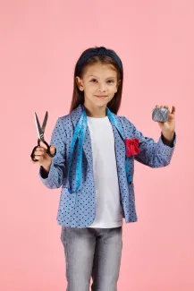 Tytöllä on toisessa kädessä sakset ja toisessa lankarulla. Kaulalla on mittanauna.