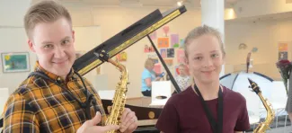 Saksofoninsoiton opettaja ja oppilas iloisena, molemmilla saksofonit käsissä ja taustalla näkyy flyygeli
