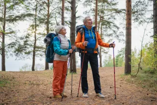 Varttuneempi nainen ja mies patikoivat luonnossa, reput selässä ja kävelysauvat kädessä.