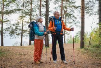 Varttuneempi nainen ja mies patikoivat luonnossa, reput selässä ja kävelysauvat kädessä.